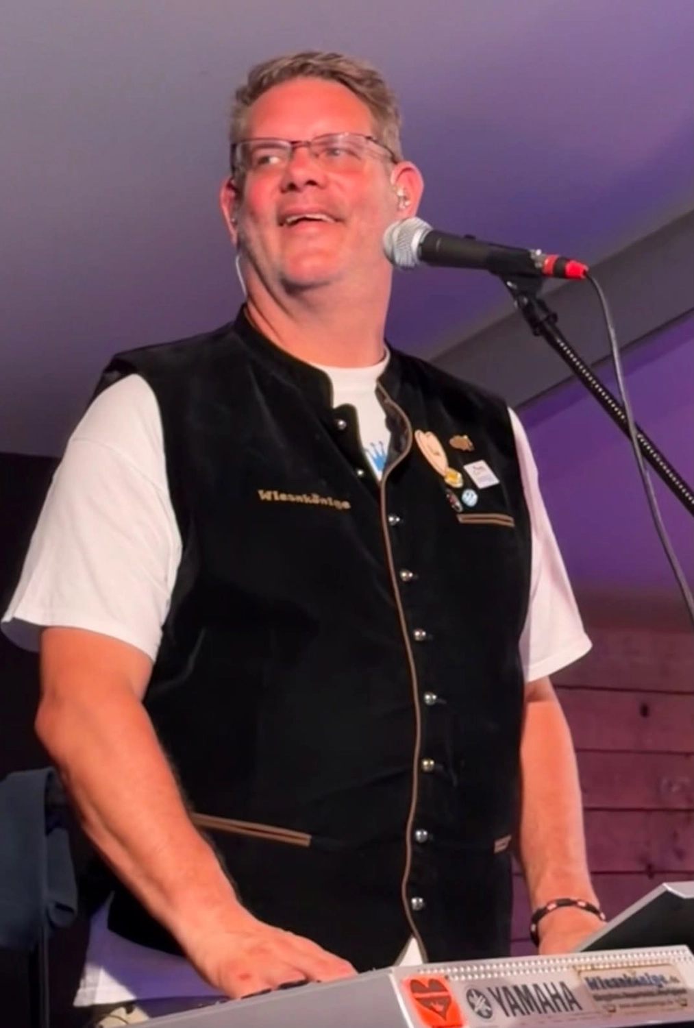Musiker in Wiesnkönige-Weste singt am Mikrofon im violetten Bühnenlicht