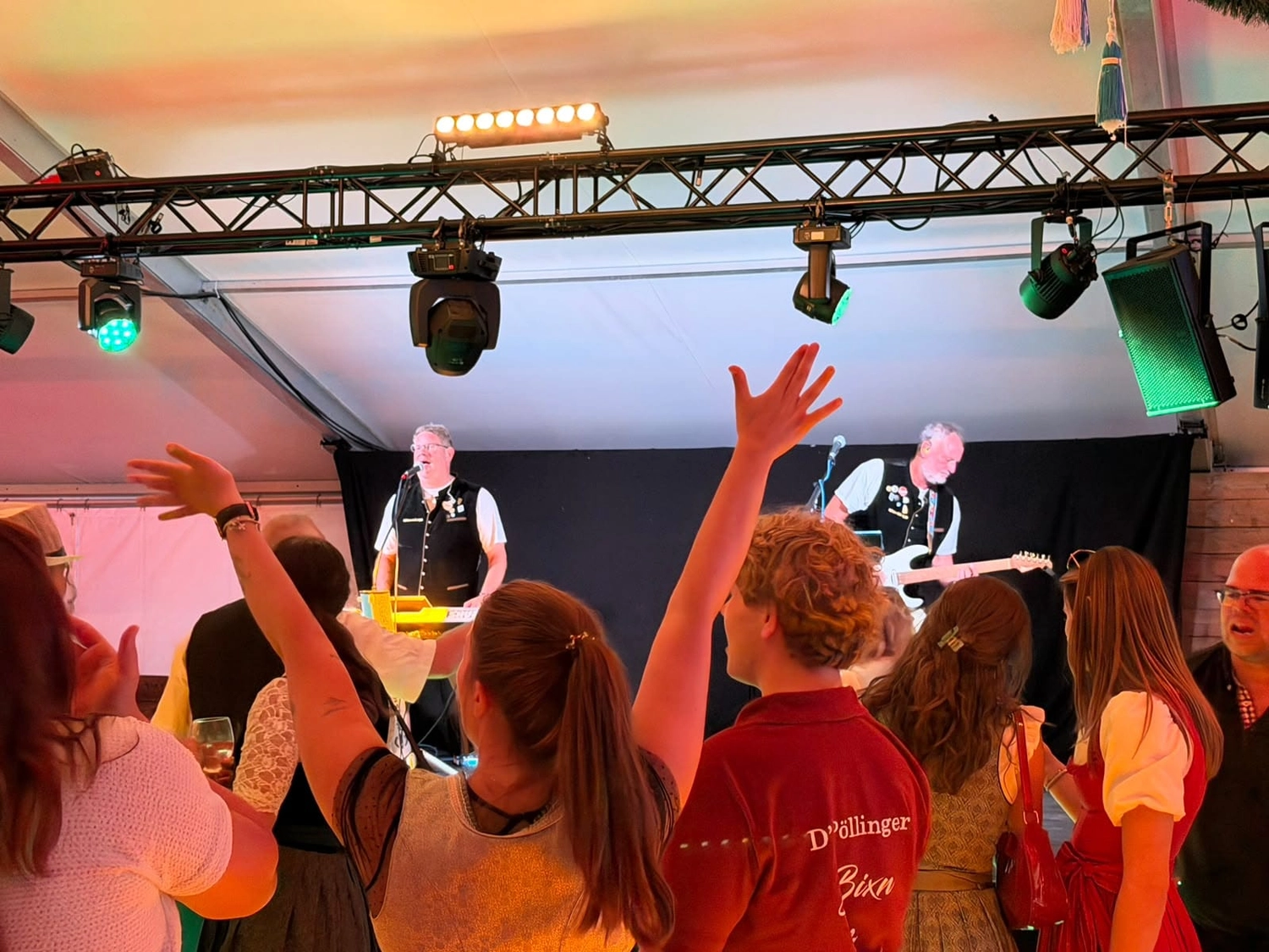 Zwei Musiker der Wiesnkönige auf der Festzeltbühne, davor feierndes Publikum mit erhobenen Händen