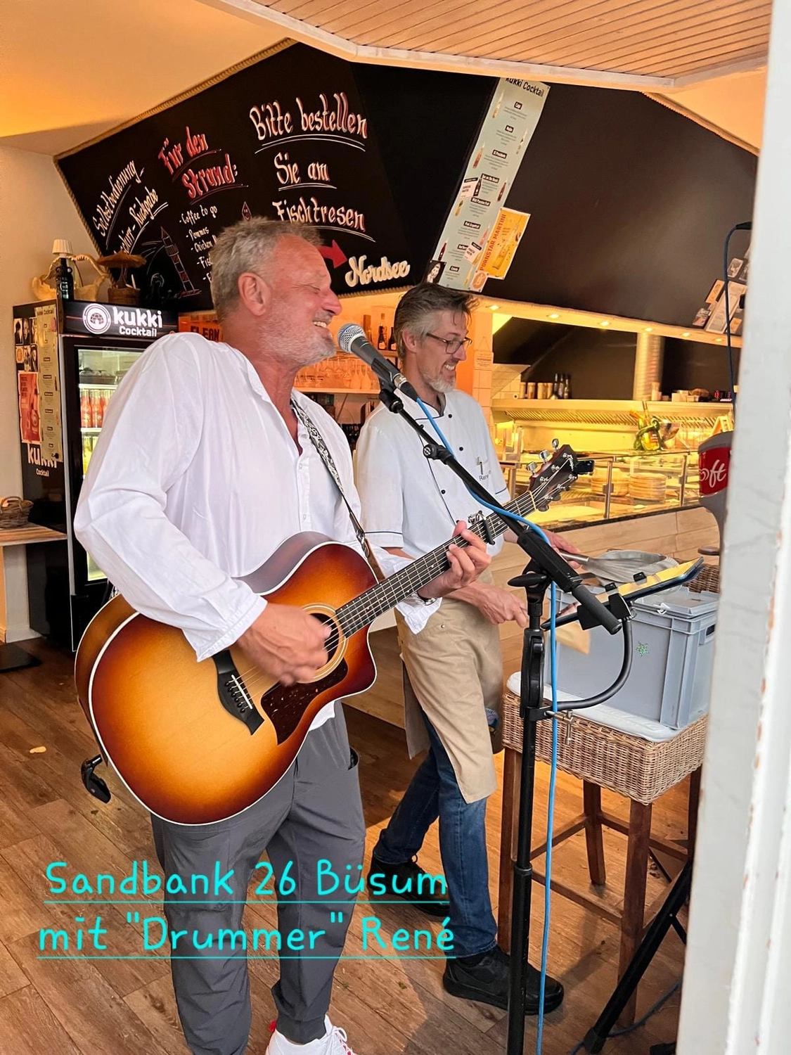 Michael Stern mit Akustikgitarre am Mikrofon in einem Bistro, daneben ein zweiter Musiker