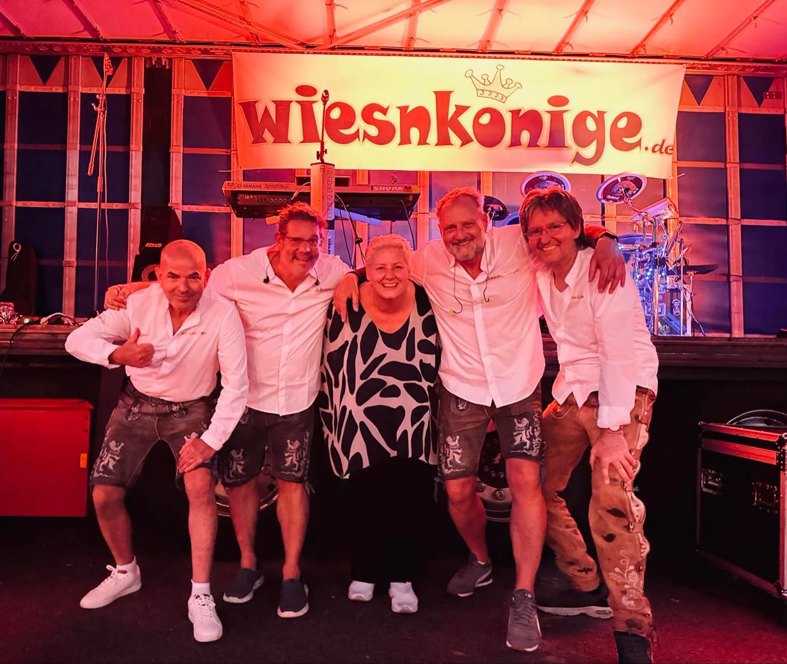 Fünfköpfige Besetzung der Wiesnkönige Arm in Arm vor dem Bühnenbanner „wiesnkonige.de“