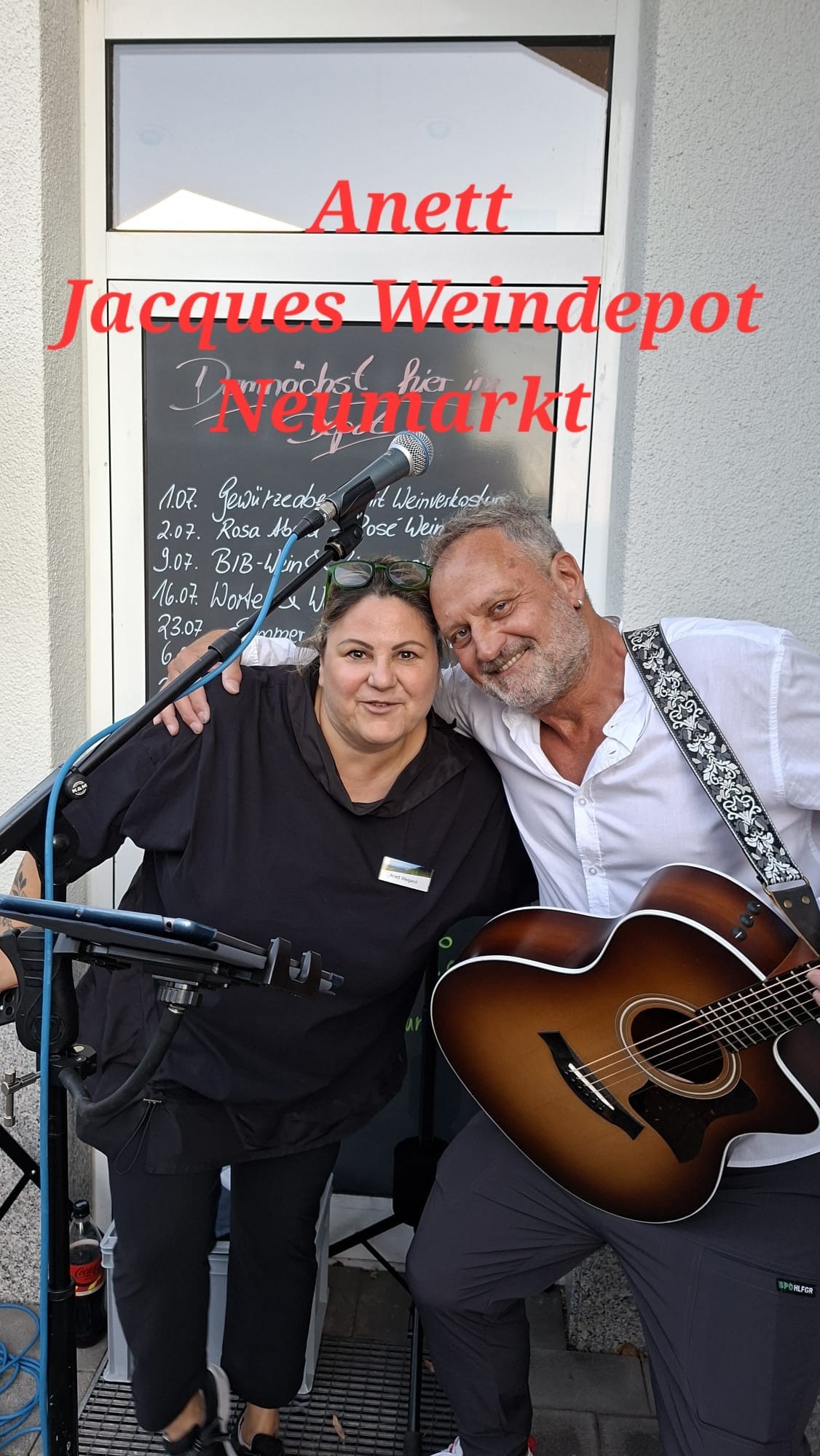 Michael Stern mit Akustikgitarre und Gastgeberin vor einer Kreidetafel, mit eingeblendetem Schriftzug „Anett Jacques Weindepot Neumarkt“