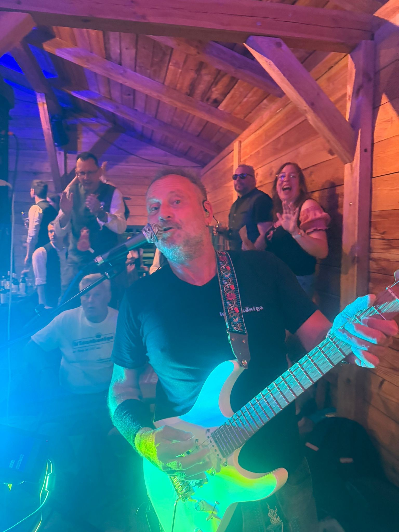 Live-Auftritt mit E-Gitarre unter Holzbalken im blauen und violetten Bühnenlicht, dahinter feiernde Gäste